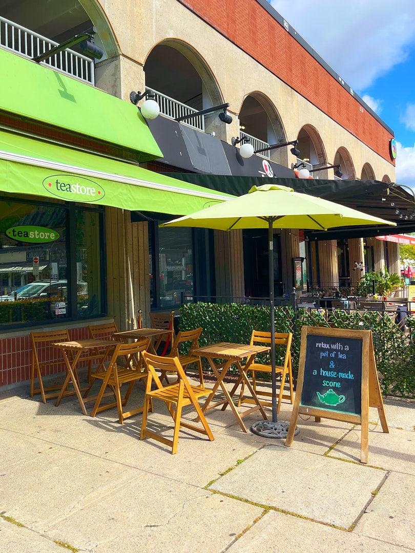 Teastore outdoor patio seating area with tables, chairs, and green umbrellas in front of a building with a green awning.